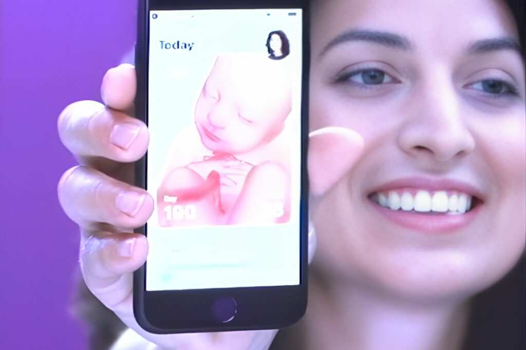 Mulher sorrindo enquanto mostra o aplicativo Pregnancy+ no celular, que exibe uma imagem 3D do bebê em desenvolvimento durante a gestação.