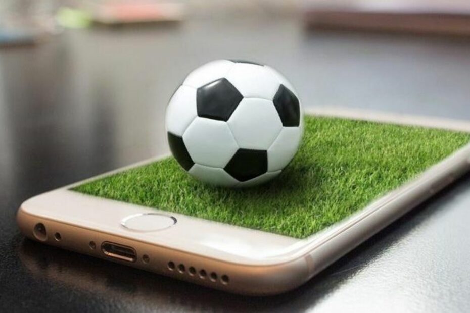 Bola de futebol sobre um smartphone com gramado virtual, representando aplicativos gratuitos para acompanhar futebol e conteúdos esportivos diretamente pelo celular.